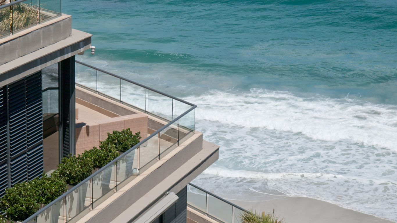 Luxury marine property overlooking the beach with clear ocean waves and modern architecture featuring glass railings and greenery.